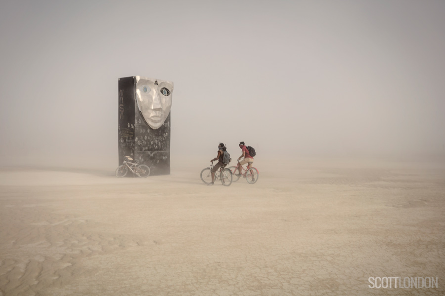 Two Burners ride by an art installation on a dusty afternoon at Burning Man 2018. (Photo by Scott London)